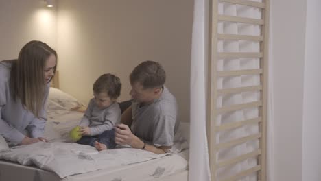 baby and his parents sitting on the bed. the baby picks up a green apple and shows it to his mother