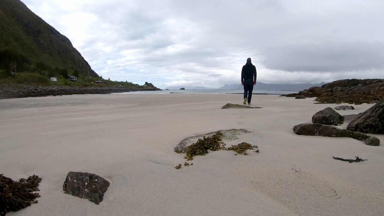 Static shot of male tourist walking across Rorvikstranda beach toward ocean under overcast sky