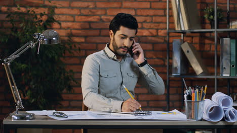 Buen-Hombre-De-Negocios-árabe-Sentado-En-Su-Escritorio-Sonriendo-Y-Hablando-Por-Teléfono