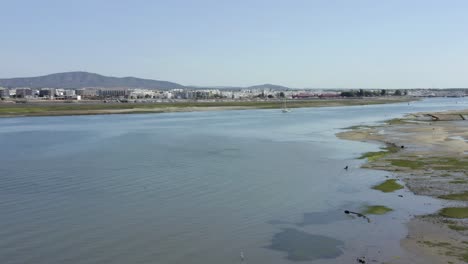 Drone-Aerial-flying-low-altitude-over-blue-river-with-sail-boat-sailing-though-calm-waters,-olhao-low-tide