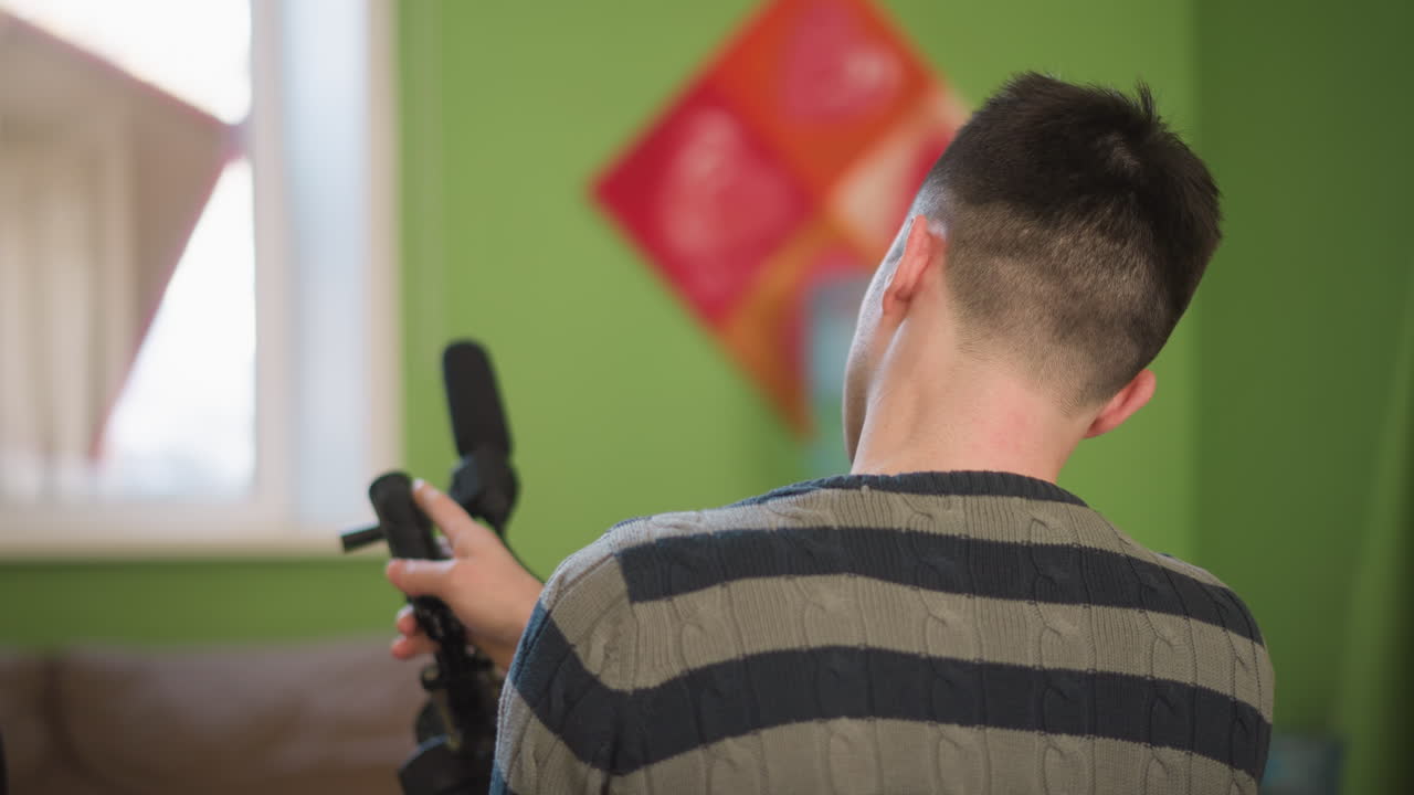Man adjusting video camera with microphone attached in creative workspace. Focused on preparing equipment for filming, surrounded by modern furniture and colorful walls, bright natural light