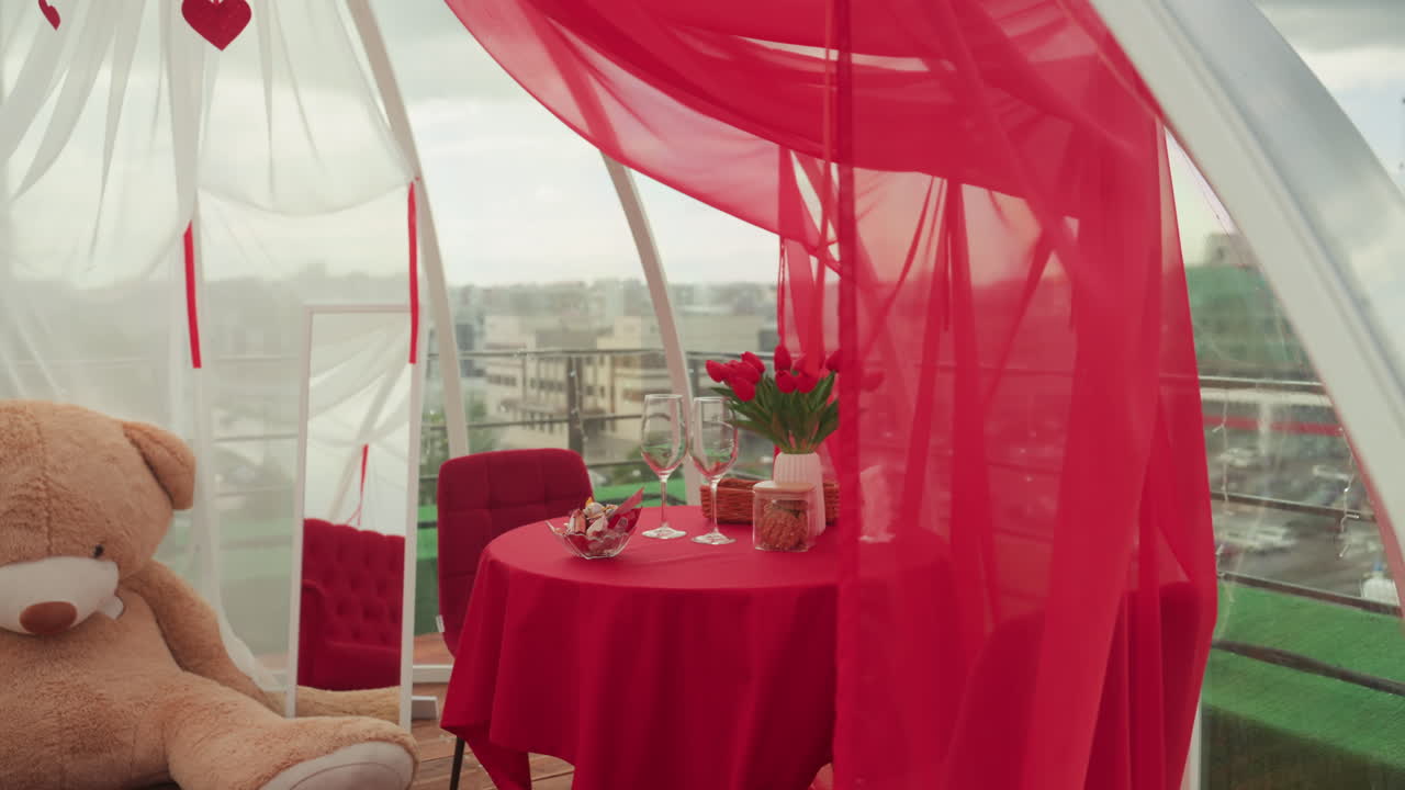 rooftop table with teddy bear romantic red drapes frame cozy intimate date setting with plush bear on chair, red tablecloth, bouquet in vase, candles, wine glasses and skyline view playful proposal