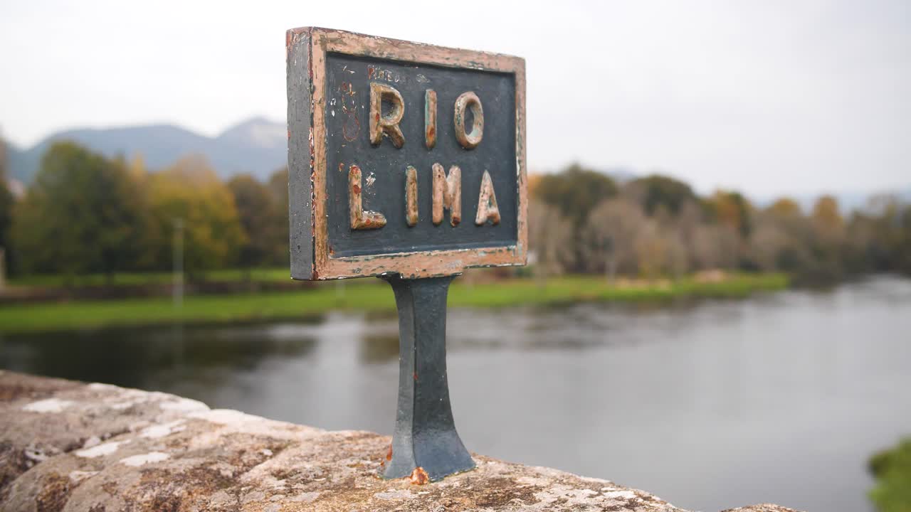 Rio Lima sign on an old stone bridge with a scenic riverside and trees in the background