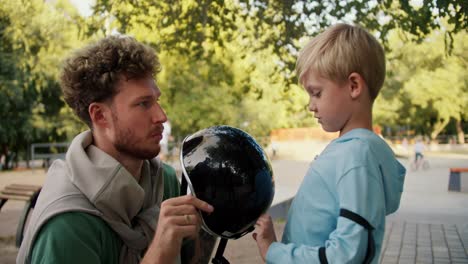 A-father-with-curly-hair-and-stubble-in-a-green-T-shirt-helps-his-son-in-a-blue-sweater-put-on-a-black-helmet-before-riding-a-skateboard-at-a-skatepark-in-the-park.-Little-blond-boy-preparing-to-ride-a-skateboard