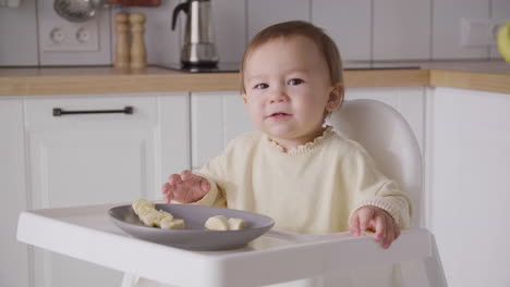 cute baby girl eating banana slices sitting in her high chair in the kitchen 2