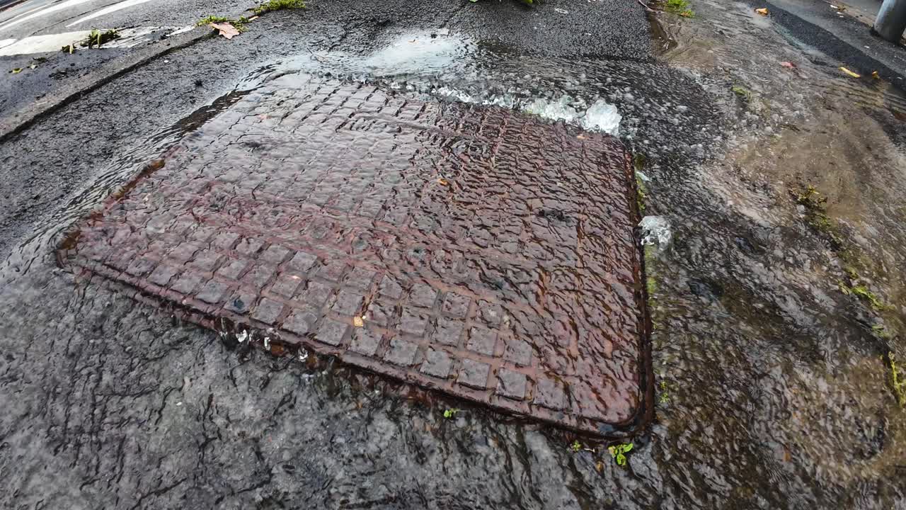 Burst Drain Cover with Treated Water Pouring Out into Street. Leaking Liquid in Need of Repair Maintenance from Water Company Professional Plumber Repairman. Wide Angle.