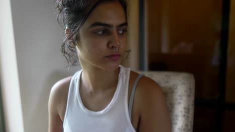 Woman-in-a-white-tank-top-looking-thoughtful-while-seated-indoors-in-a-cozy-room