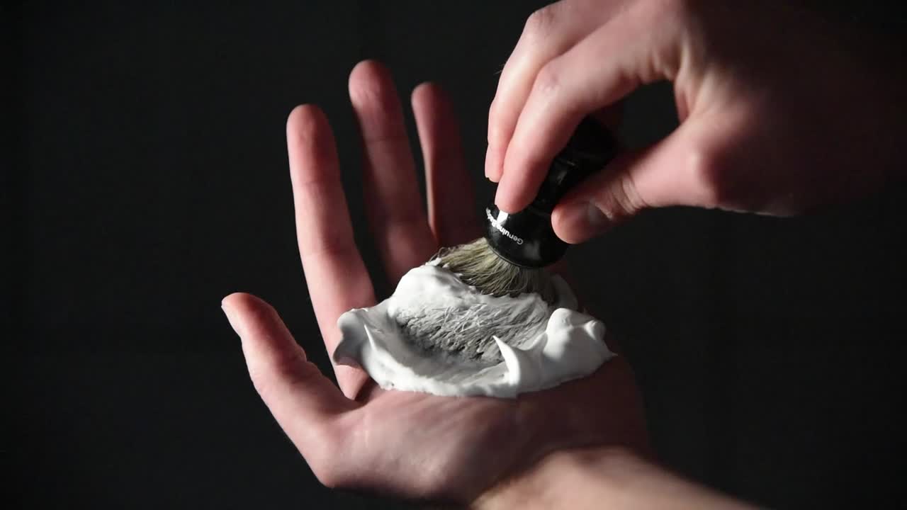 Nærbillede af kaukasisk hånd, der skummer barberskum op på en sort barberkost. Mørk indendørs baggrund