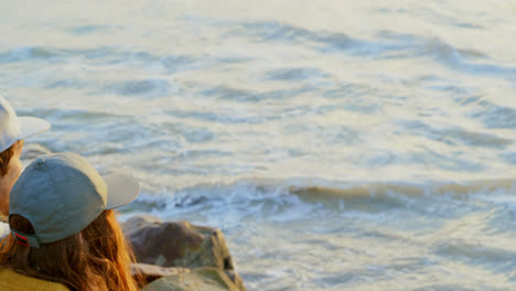 Rear-view-of-young-caucasian-couple-sitting-on-rock-and-looking-at-sea-on-the-beach-4k