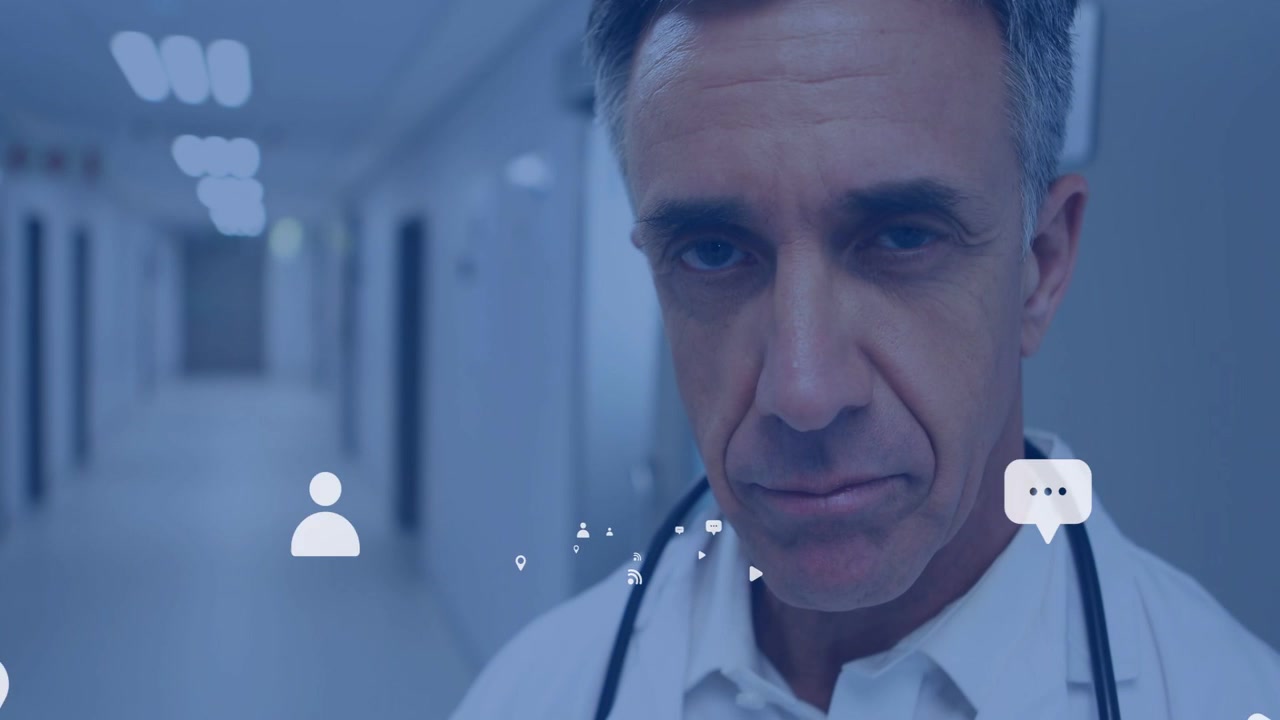 Medico con stetoscopio in piedi in corridoio, infermiera in divisa blu al telefono che attiva icone, lui che alza lo sguardo per prestare cure.