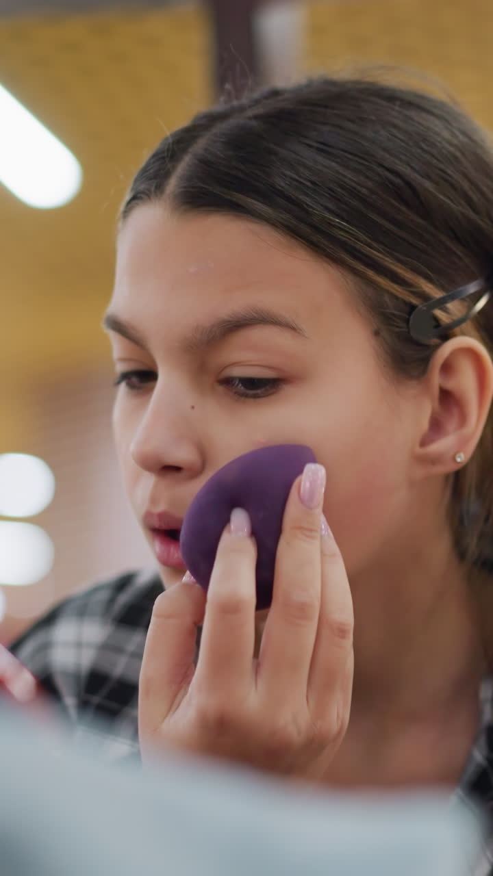 adolescente caucásica aplicándose maquillaje con una esponja, primer plano del rostro con expresión neutra y pelo recogido, difuminado preciso de la base con una esponja morada bajo luces fluorescentes, ambiente instructivo y tranquilo