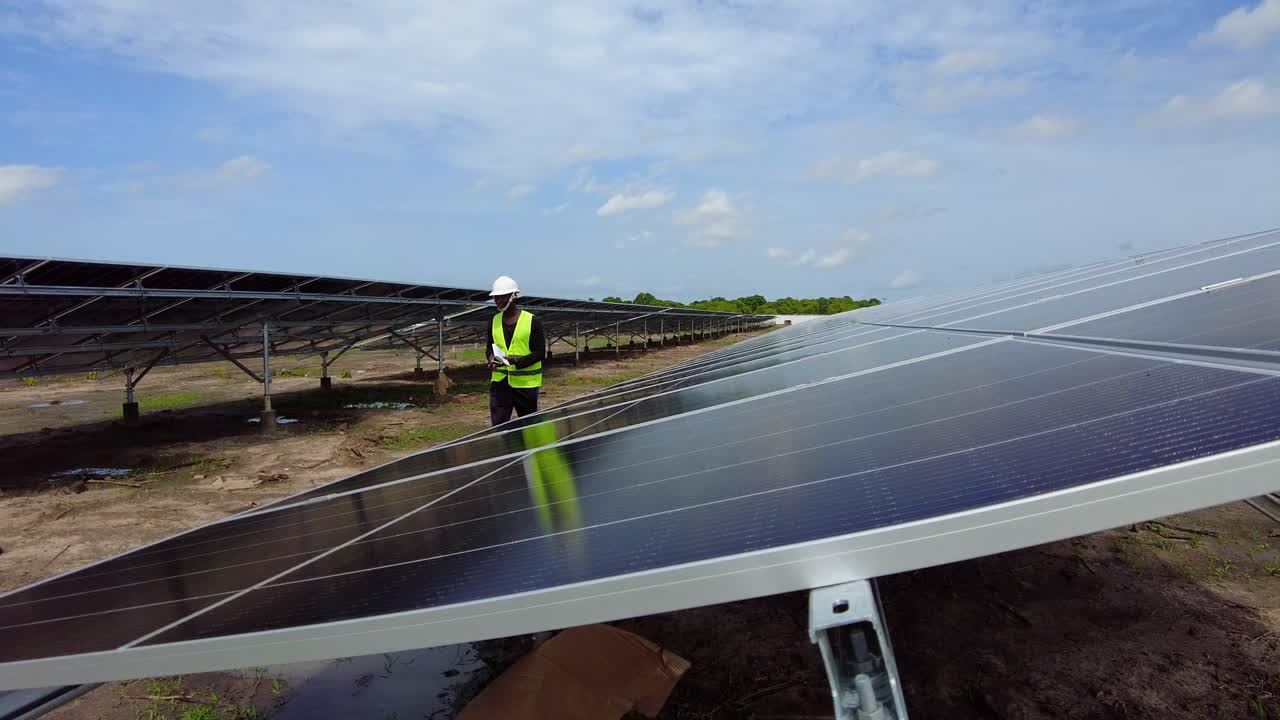 ingeniero africano negro caminando en una granja de energía renovable de paneles solares con casco protector llevando un cuaderno de papel con datos de mediciones de la eficiencia de la planta fotovoltaica en áfrica