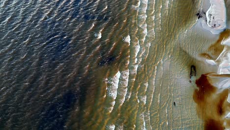 Vista-Aérea-De-Una-Playa-Con-Vetas-De-Tierra-Resbaladiza-Visibles-En-La-Superficie-Del-Agua,-Restos-De-Barcos-Antiguos,-Junto-Con-Olas-Rompientes-Y-Arena-Iluminada-Por-El-Sol.