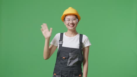 asian woman worker wearing goggles and safety helmet smiling, waving hand, and saying bye while standing in the green screen background studio