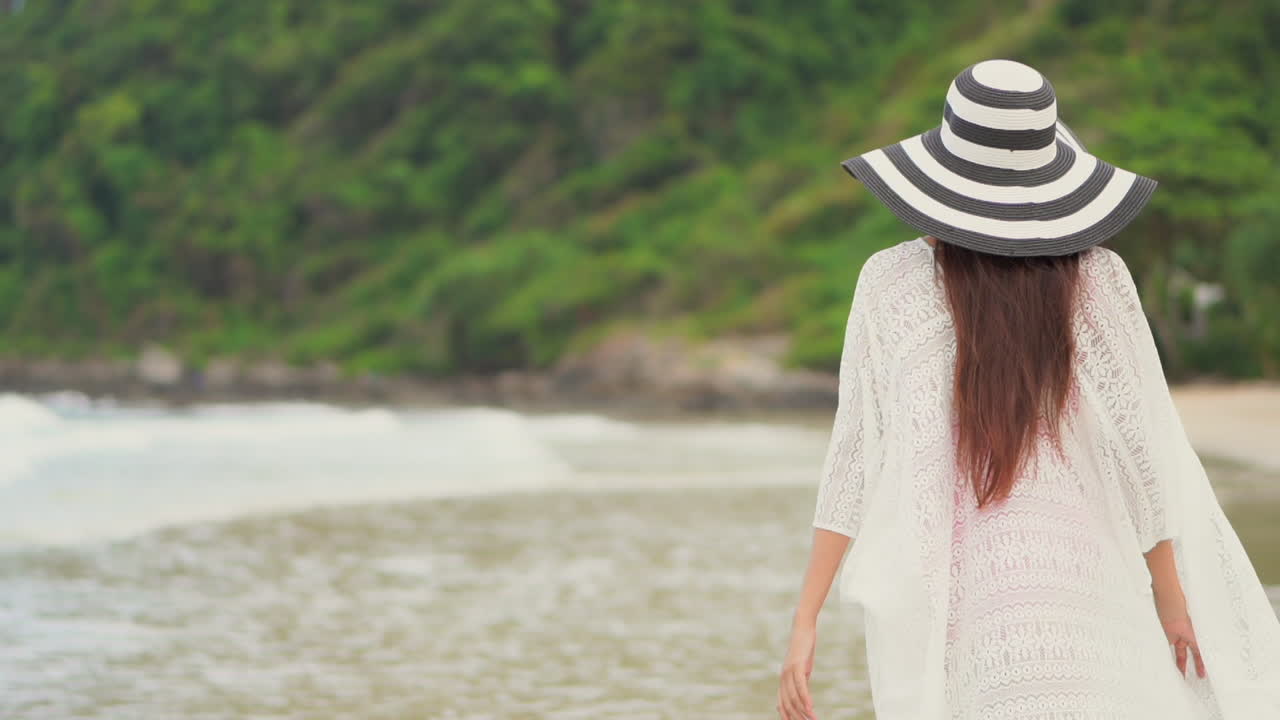 하얀 드레스와 모자를 쓴 여성의 뒷모습이 해변에서 맨발로 걷는다
