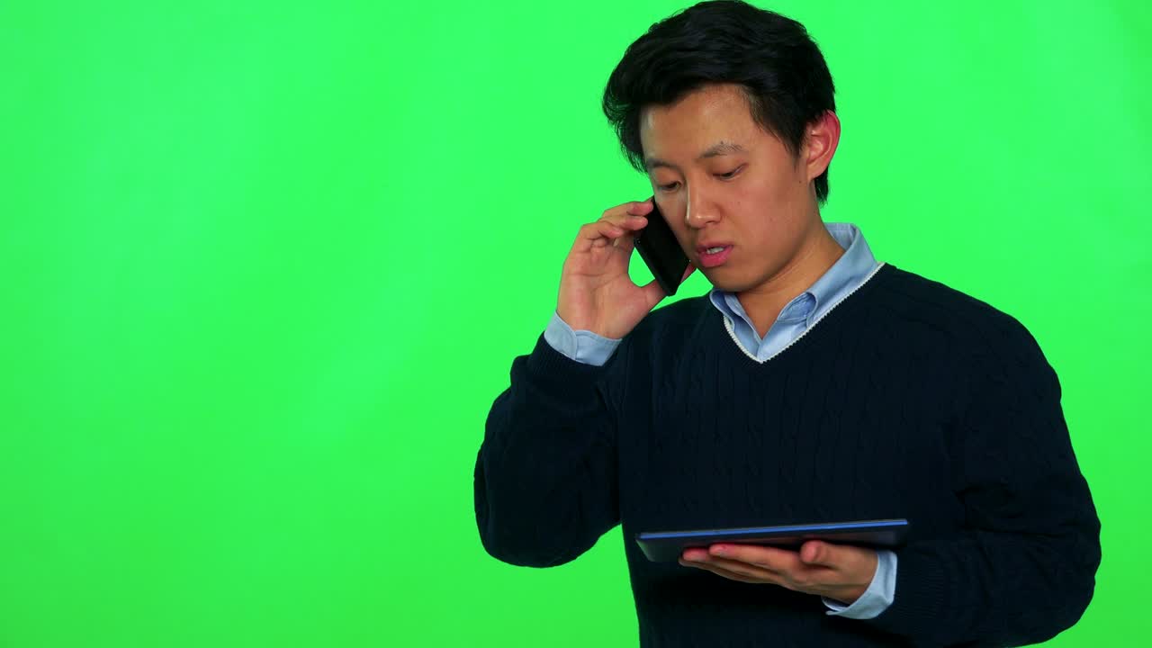 젊은 아시아 남자가 태블릿을 들고 보고 스마트폰으로 대화하고 있습니다. - 그린 스크린 스튜디오