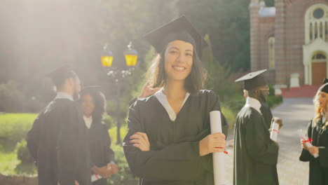 retrato de una encantadora graduada sonriente con ropa tradicional y gorra mirando a la cámara con su diploma en manos cruzadas