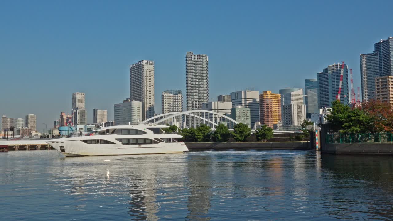 A sleek white luxury yacht cruises on the calm water of Tokyo Bay, with modern city buildings and a bridge visible in the distance under a clear blue sky.
