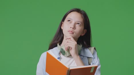 close up of asian teen girl student with a backpack reading book and thinking and looking around then raising her index finger while standing in the green screen background studio