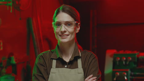 caucasian young woman welder wearing apron and goggles looking at camera with arms crossed in workshop