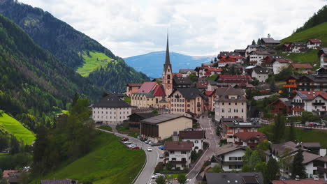 drone footage of a slow approach towards an alpine village with a tall church steeple, surrounded by green mountain slopes and valleys in the dolomites, italy