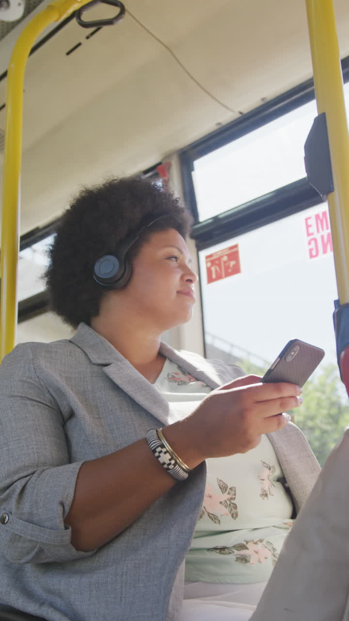 video vertical de una mujer biracial feliz con auriculares usando un teléfono inteligente en el autobús