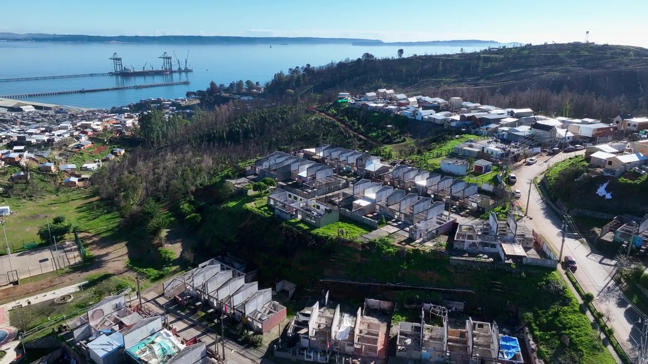 Cirkulær flyvning omkring et kvarter under genopbygning med nye tage og byggestel, der afslører den industrielle mole, en rolig bugt og en fjern kystlinje. Lirquen, Penco, Chile