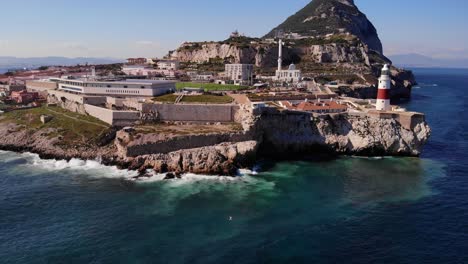 Wellen-Schlagen-Auf-Die-Felsen-Unterhalb-Des-Leuchtturms-Am-Europa-Point,-Während-Weiße-Möwen-Mit-Dem-Berühmten-Felsen-Von-Gibraltar-Umherfliegen-Und-Schiffe-In-Der-Bucht-Von-Algeciras-Ankern