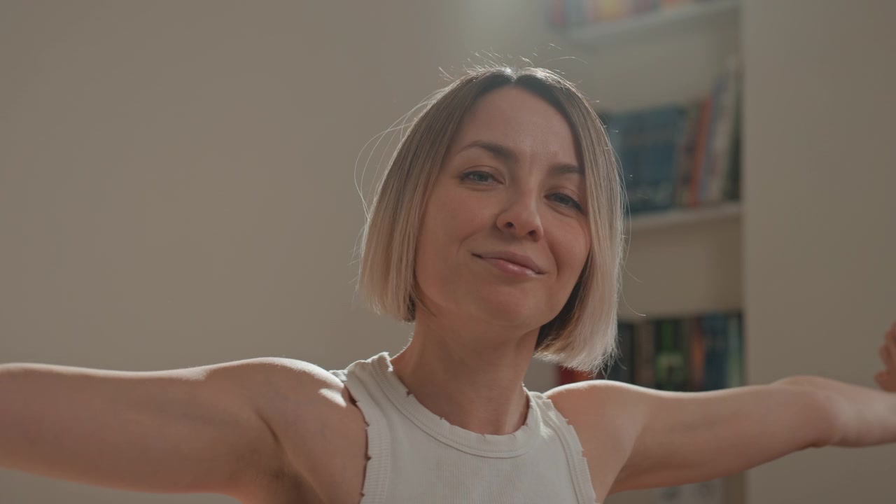 Mujer caucásica sonriente disfrutando de la música, estirándose y bailando en un dormitorio soleado. Viste una camiseta sin mangas blanca, con cabello rubio corto tipo bob y una expresión relajada y segura. Retrato de primer plano, con estantería y lámpara.