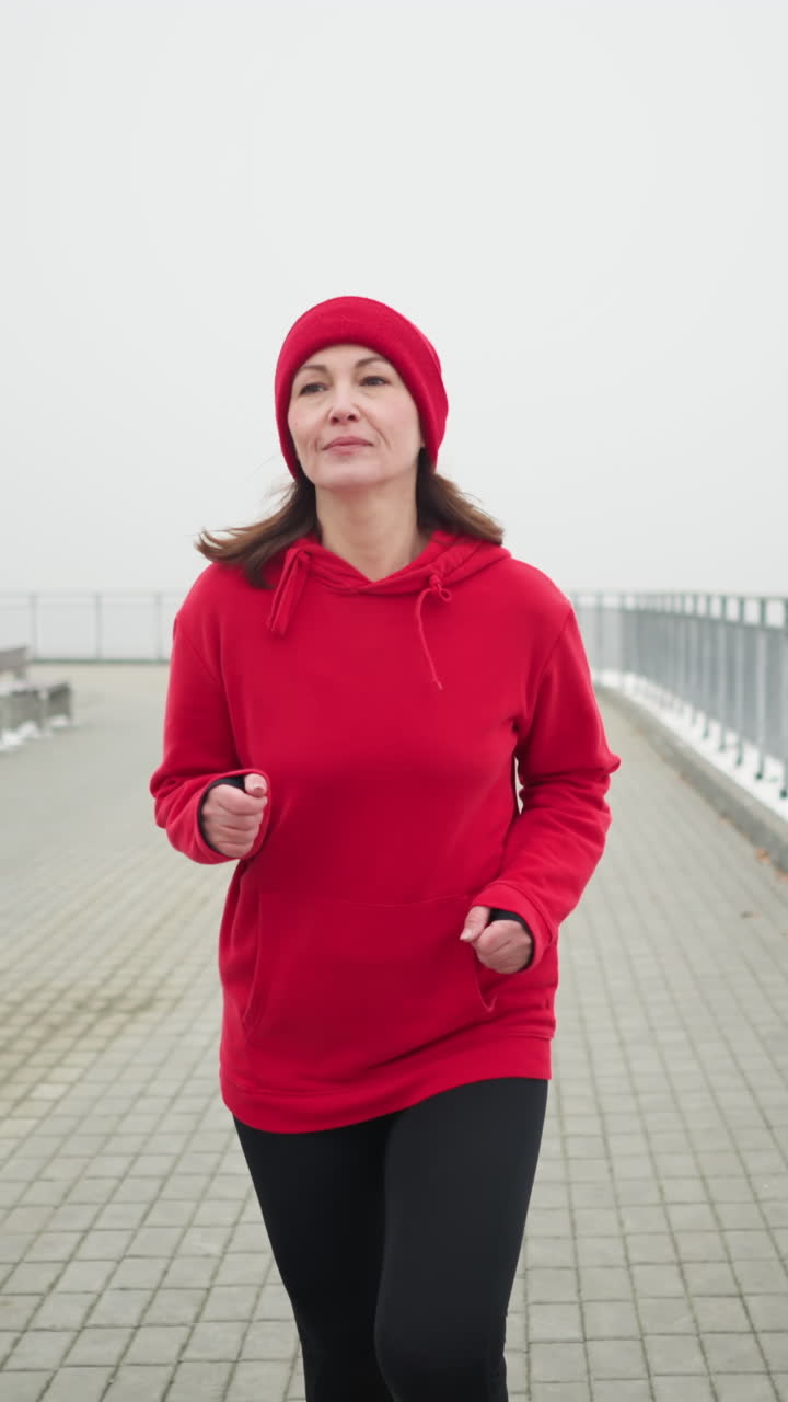 middle aged woman in red beanie and jacket jogging near iron railing on snowy pathway surrounded by benches in foggy serene winter park showcasing cold weather fitness routine with breath visible
