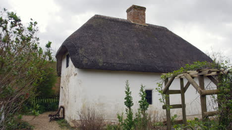 Thatched-roof-cottage-with-chimney,-surrounded-by-lush-greenery-and-a-wooden-trellis,-evokes-rural-charm