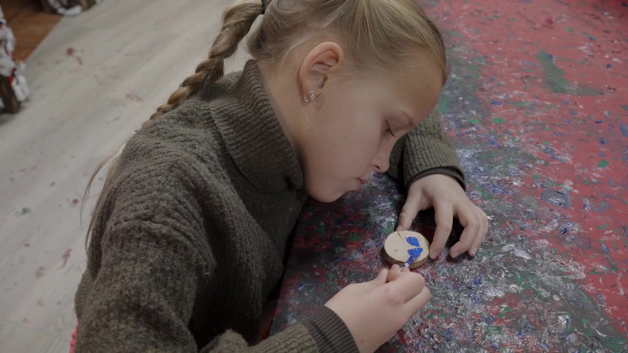 Una giovane ragazza bionda con i capelli intrecciati dipinge con cura un ornamento in legno a forma di viso di Babbo Natale con un pennello a un tavolo da laboratorio colorato e schizzato di vernice