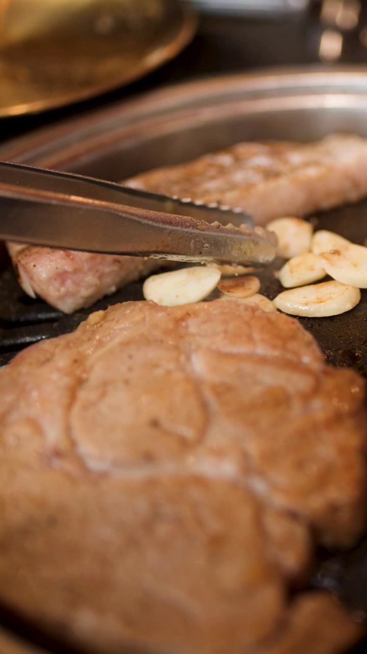 暖かく自然な照明の下、卓上韓国焼肉で焼いている豚肉に、トングでニンニクスライスを加える。