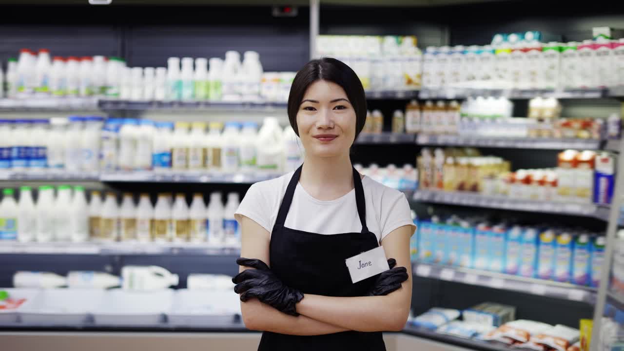 乳製品の棚があるスーパーマーケットに立つ、黒いエプロンを着た魅力的な若いアジア人女性店員のポートレート。