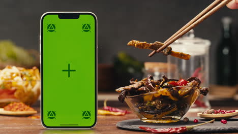 a green screen mobile phone, a portable communications device, is on a table next to a bowl of canned corn. the product is a useful gadget for telephony and communication. fermented food served on table, mini corn, kimchi, eggplants, yellow cabbage