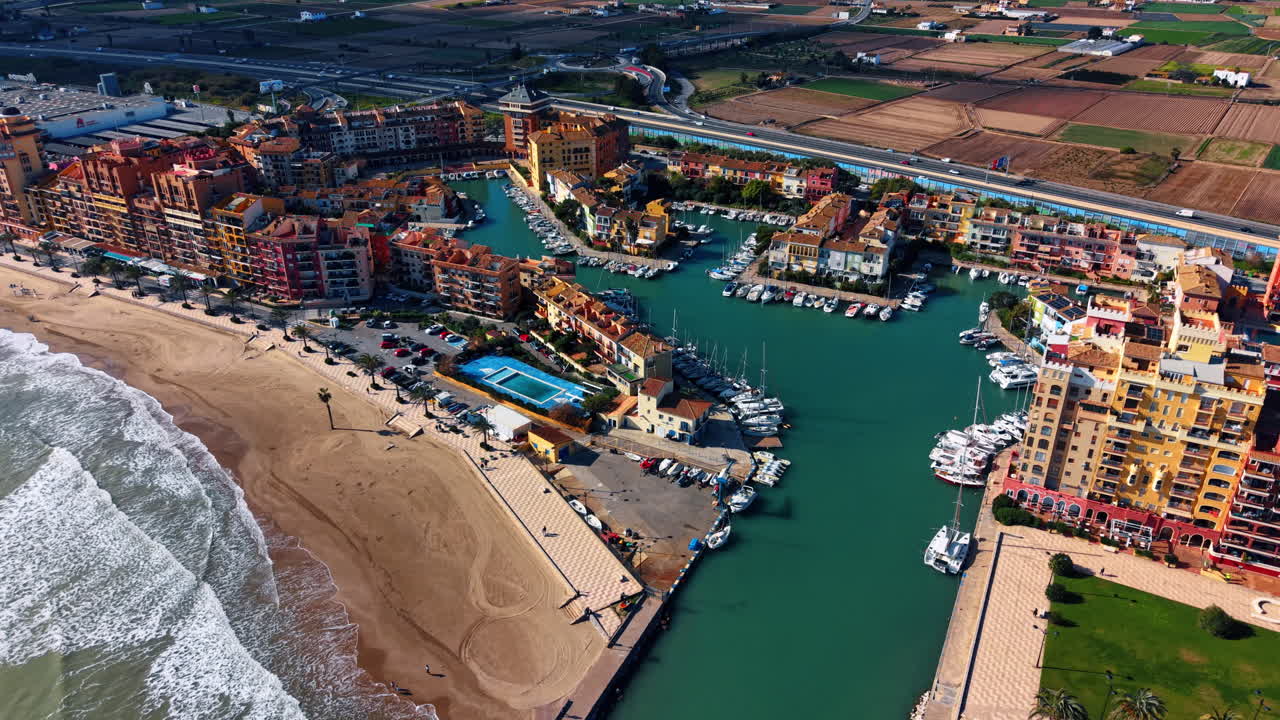 Ampia veduta aerea del porto turistico e della spiaggia di Port Saplaya. Panorama di una colorata cittadina costiera, spiaggia sabbiosa, porto per barche e campi agricoli sotto un cielo soleggiato