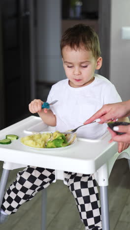 baby boy sits at high chair waving his feet. toddler eats from fork. mom uses fork to mash the food.