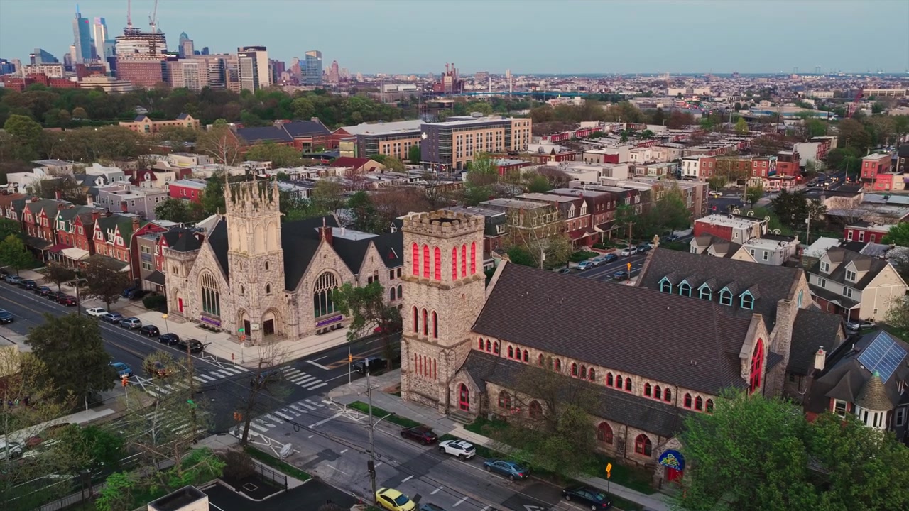 Veduta aerea con drone di due chiese storiche in pietra fianco a fianco in un quartiere di Filadelfia al tramonto, con lo skyline di Center City che risplende in lontananza.