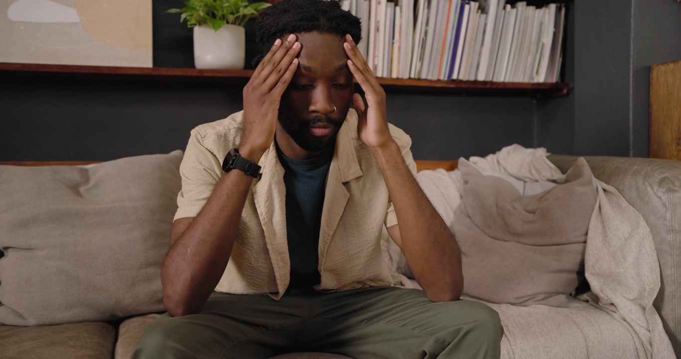 Man shifting hands then rubbing temples, leaning forward on couch near bookshelf, showing stress