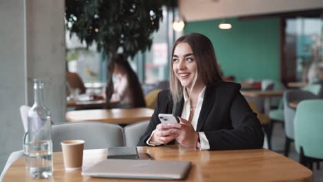 En-Un-Café,-Una-Joven-Hermosa-Vestida-De-Negocios-Sonríe-Mientras-Envía-Mensajes-De-Texto-En-Su-Teléfono-Inteligente,-Participa-En-Las-Redes-Sociales-Y-Envía-Mensajes