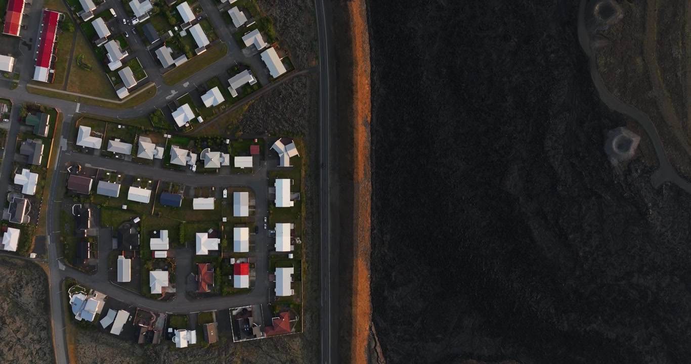 Top-down drone view of Grindavik, Iceland, contrasting a compact residential quarter with a long orange defense berm and an expansive hardened lava field.