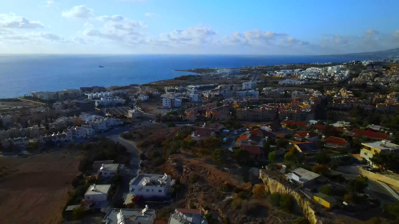 Aerial drone view of coastal suburb with modern buildings, open land, and calm sea in background under clear sky, showing peaceful urban sprawl near waterfront from above