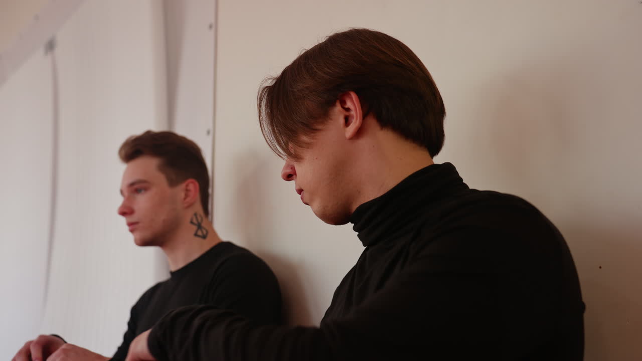 Man in black uniform walks to sit another man near punching bag with focused profile view, showing concentration, calmness, discipline, and determination in training environment with natural light