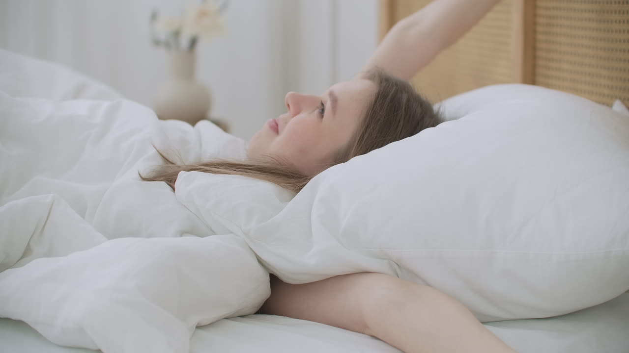 la hermosa morena se despierta por la mañana se extiende en la cama el sol brilla sobre ella desde la gran ventana una joven feliz saluda el nuevo día con una cálida llamarada de luz solar.