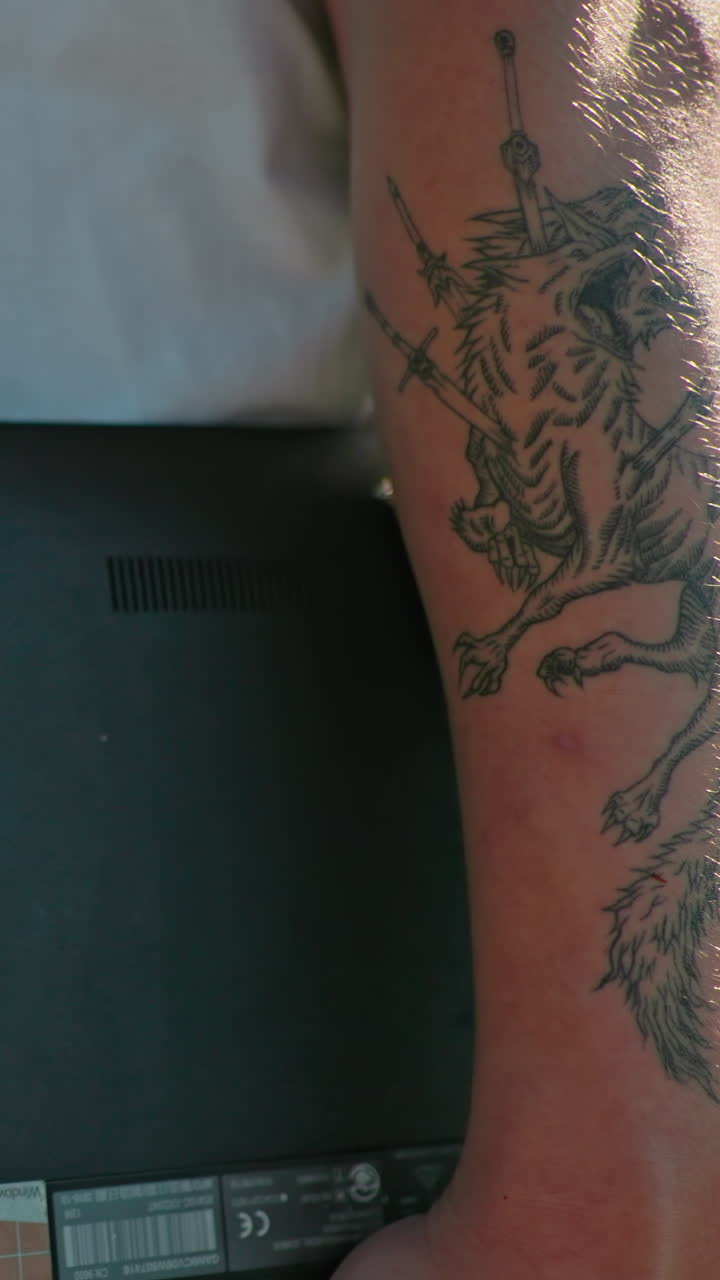 핑크 을 입은 사람과 함께 는 동안 노트북을 들고 있는 문신한 팔의 클로즈업, 밝은 빛 아래 문신 예술의 캐주얼 상호 작용과 세부 사항을 포착합니다.