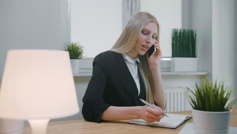 business woman talking on smartphone in office. elegant young blond female in office suit sitting at workplace and negotiating via mobile phone in hand writing down necessary information into notebook