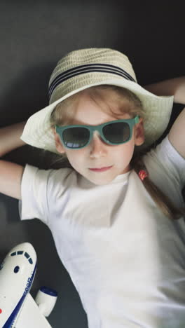 small girl in straw hat with hands behind head relaxes at home before departure. upset preschooler in sunglasses prepared for vacation upper closeup