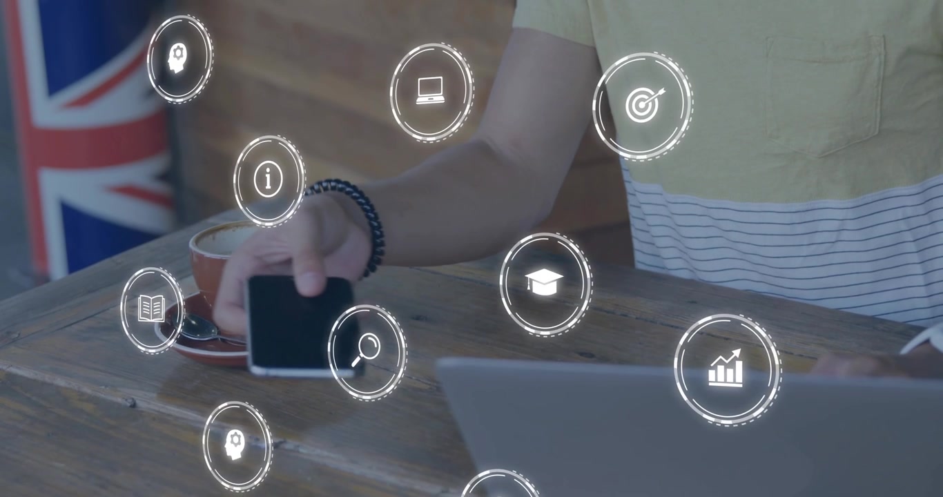 Man typing on laptop causing education icons to swirl around smartphone and coffee on cafe table