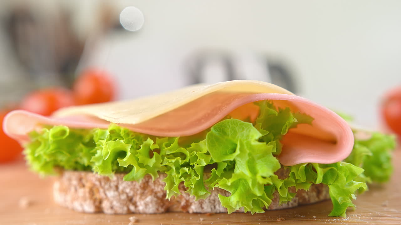pan de trigo con lechuga, jamón y rebanada de queso cayendo a cámara lenta en un sándwich en una tabla de cortar de madera en una cocina