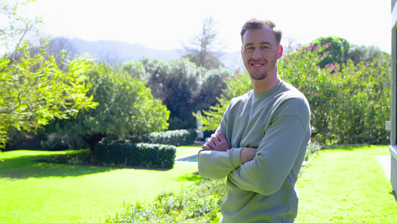 Man entering sunlit garden, crossing arms, turning toward camera, greeting with warm smile
